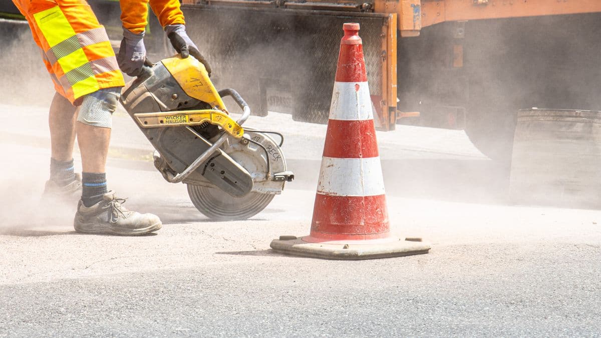 A construction worker in high-visibility gear cutting into the ground with a gas-powered saw beside a traffic cone, dust rising around the work area