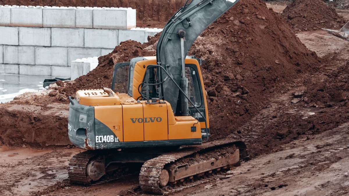 An excavator grading a hillside construction site next to a retaining wall, with dry Mediterranean-style trees in the background