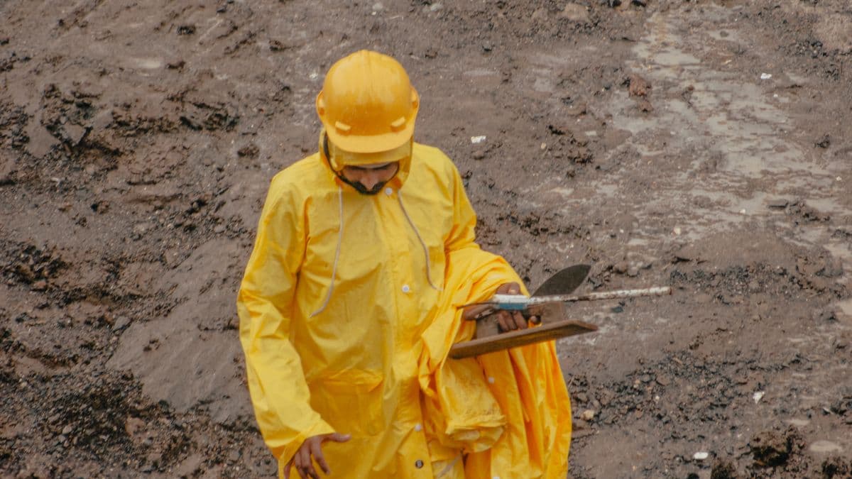 A worker in yellow rain gear walks a muddy, rain-soaked construction site holding a tablet during a wet-weather inspection.