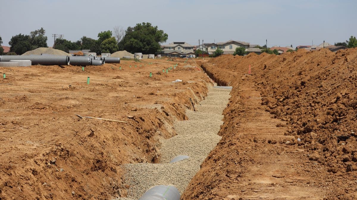 An open storm drainage trench with gravel bedding and pipe cut through flat Texas soil, subdivision houses visible in the distance